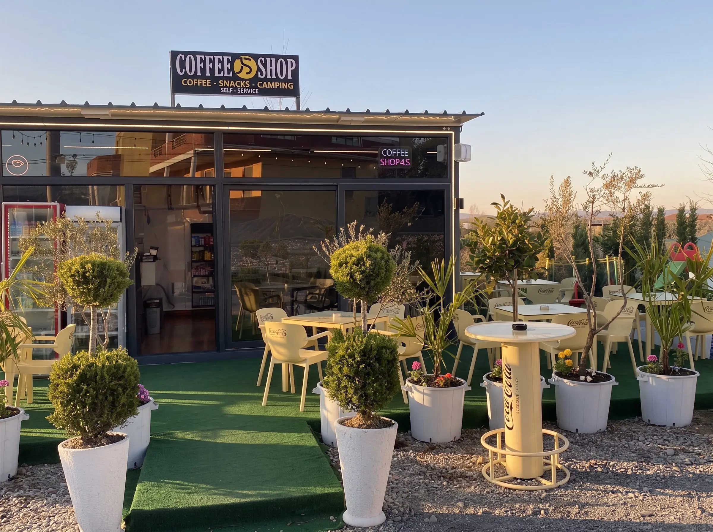 Coffee Shop JS in late-afternoon sun: the terrace on green turf, small trees in white pots, high tables