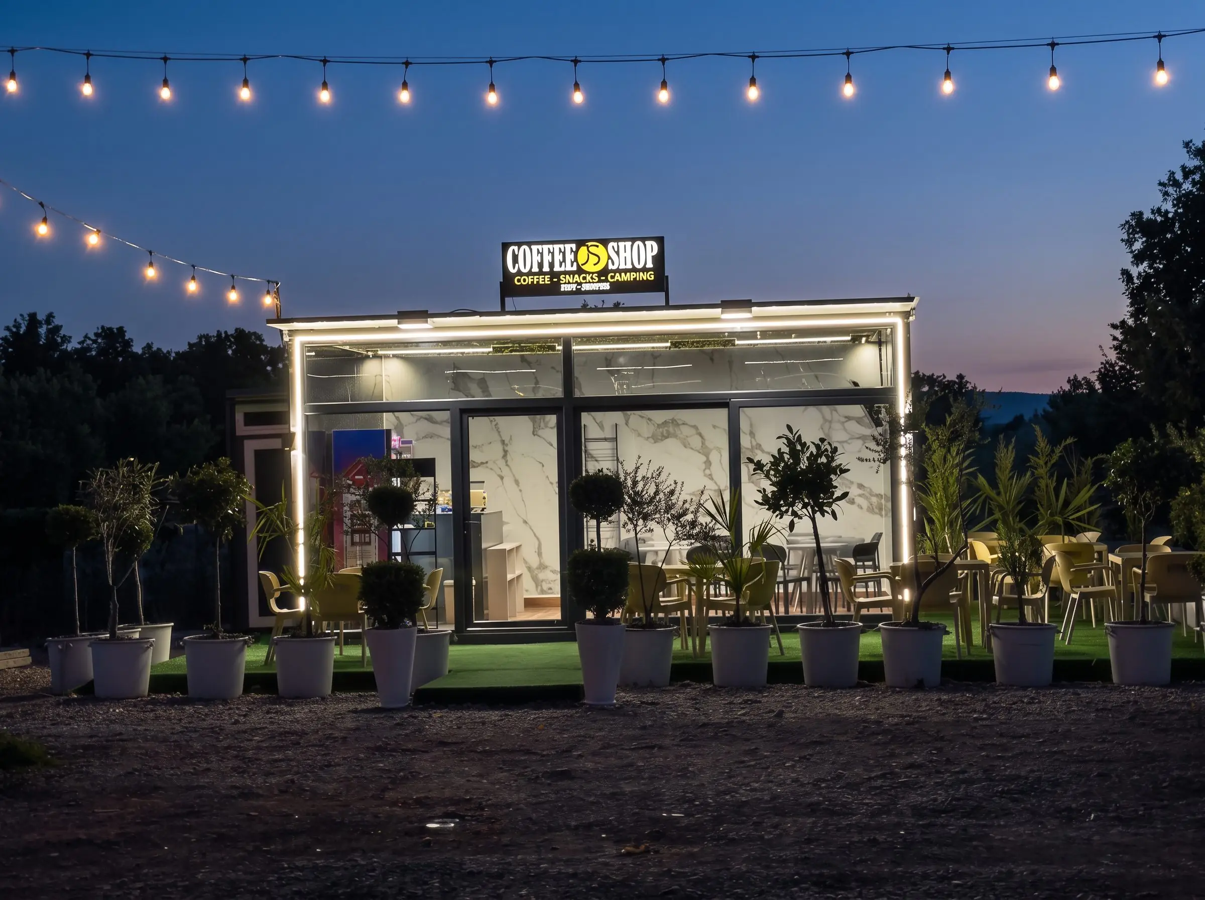 Coffee Shop JS at blue hour, its sign lit, under a line of warm string lights