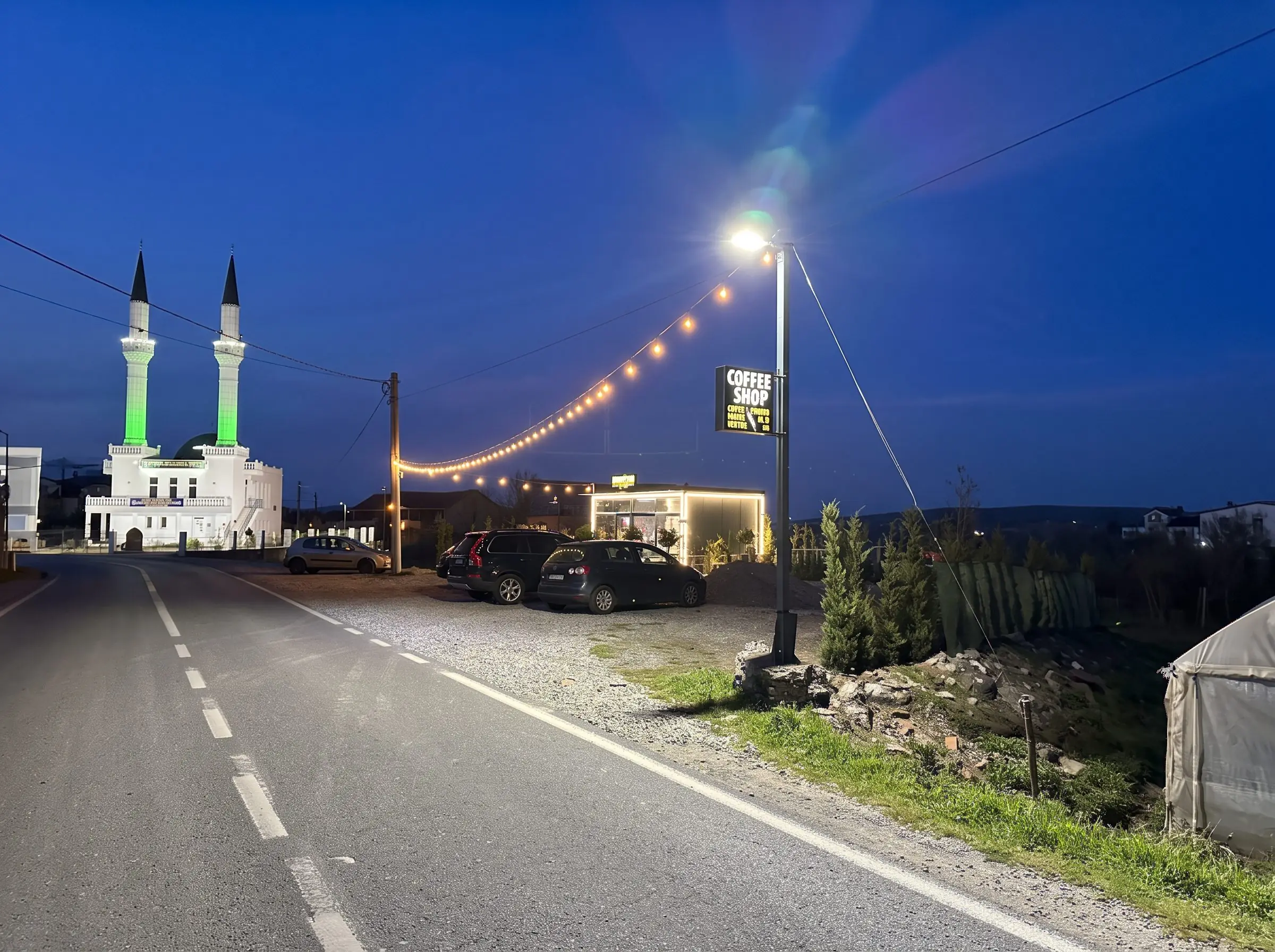 The road at blue hour: the mosque's green-lit minarets, a street lamp and the string lights of Coffee Shop JS by the roadside